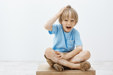 Indoor shot of unhappy cute blond child with vitiligo, having two-colored skin, sitting on floor with crossed feet, touching head and screaming, being displeased and disobedient over gray wall