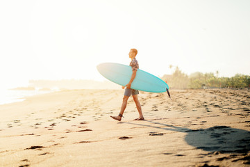 Wall Mural - Portrait of happy surfer in hawaiian t-shirt walking with surf board on the beach at sunset