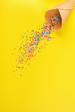 Ice Cream Cone And Colorful Spilled Sprinkles On Yellow Background. Concept, Minimalist, High Angle, Vibrant Colors.