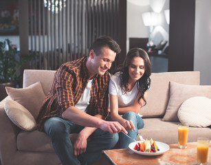 Lets taste this. Portrait of hungry young man and woman are eating sweet dessert by forks. They are looking at food with appetite and smiling while sitting on sofa at home 