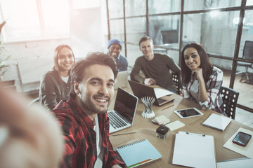 Joyful day. Portrait of successful young people are sitting together at table with smart devices and taking selfie photograph while being in friendly mood. They are smiling and expressing gladness