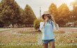 © skyNext - Smiling black tourist girl in the hat is standing outdoors and holding her vintage film camera near her face prepared to take photos, with a huge flowerbed behind and copy space area on the left