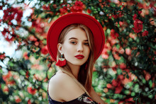 Outdoor Portrait Of Young Beautiful Girl With Red Lips, Long Blonde Hair, Wearing Hat, Long Tassel Earrings, Cold Shoulder Dress, Posing Near Blooming Tree. Summer Fashion Concept. Copy Space 