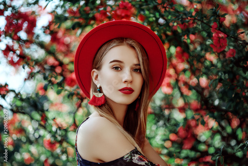 Fotomural Outdoor portrait of young beautiful girl with red lips, long blonde hair, wearing hat, long tassel earrings, cold shoulder dress, posing near blooming tree