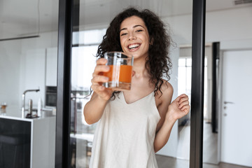 Poster - Photo of gorgeous brunette curly woman wearing silk leisure clothing smiling, while showing glass of orange juice at camera