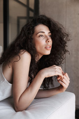 Wall Mural - Closeup photo of dreaming tender woman 20s with long curly hair wearing silk leisure clothing looking aside with brooding glance, while lying on sofa