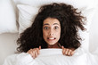 Photo from above of amusing uptight woman 20s with dark curly hair grimacing at camera, lying in bed under white blanket