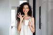 © Drobot Dean - Portrait of caucasian curly woman with long dark hair smiling at camera, and showing ok sign, while drinking tea at hotel apartment or flat in morning