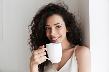 Poster - Portrait closeup of gorgeous happy woman with long curly hair smiling at camera, and drinking tea from cup in morning at hotel apartment