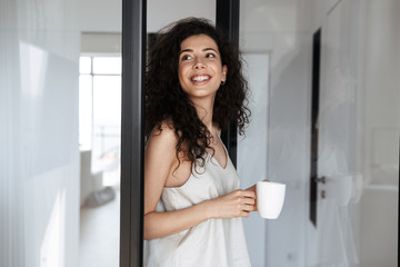 Poster - Photo of adorable curly woman with long dark hair smiling and looking aside, while standing near glass door in house with cup of tea