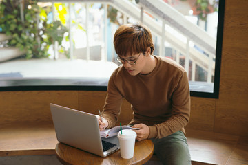 Cheerful male journalist in trendy glasses happy to finishing work on book review rereading written in notebook text before calling to editor to present publication drinking coffee for lunch in cafe