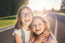 Two Joyfull Girls Standing On The Road, Hugging And Smiling Against The Sunset. Sincere Emotion. Girls Without Front Teeth.