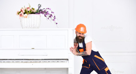 Wall Mural - Man with beard worker in helmet and overalls pushes, efforts to move piano, white background. Heavy loads concept. Loader moves piano instrument. Courier delivers furniture, move out, relocation.