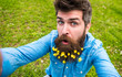 Natural beauty concept. Hipster on surprised face sits on grass, defocused. Guy with lesser celandine flowers in beard taking selfie photo. Man with beard enjoys spring, green meadow background.