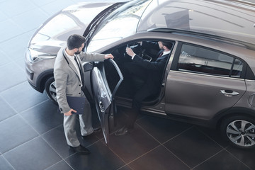 Full length high angle view at bearded car salesman opening door for customer getting into his brand new luxury car, copy space