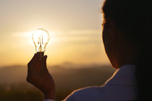 Portrait Of A Woman (girl) Holding A Light Bulb With The Sunset In The Backgroud. Concept: Energy, Technology, Power