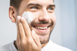 © denis_vermenko - Skin care. Handsome young shirtless man applying cream at his face and looking at himself with smile while standing over gray background and looking at camera. Close-Up. Space for text.