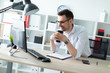 © Ivan Traimak - A young man in glasses stands near a table in the office holding a glass of coffee.