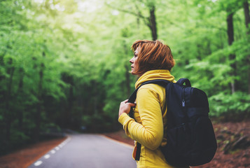 Wall Mural - tourist traveler with backpack into road at summer green forest, view back girl hiker in yellow hoody looking and enjoying the breath of fresh clean air in trip, relax holiday concept