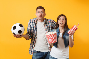 Young fun couple, woman man, football fans holding soccer ball, bucket of popcorn, plastic glass of cola, cheer up support team, isolated on yellow background. Sport family leisure lifestyle concept.