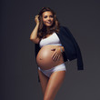 Full body portrait of attractive smiling pregnant brunette woman with slightly curly hair, wearing white sports underwear, trainers and with black jacket draped over shoulders, standing and posing.