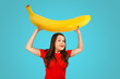 Cute funny young woman holds over herself huge yellow ripe banana on a blue background