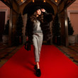 © Dmitry Tsvetkov - Gorgeous young woman with long brunette hair dressed in gray jumper, trousers and luxurious fur coat, holding handbag, posing on red carpet against building entrance and potted trees on background.