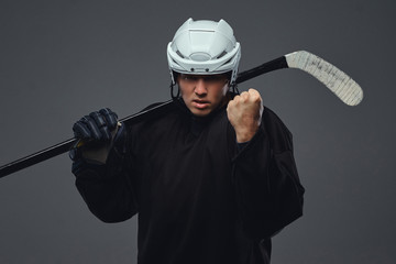 Angry professional hockey player in black sportswear standing with a hockey stick on a gray background.