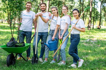 Wall Mural - young volunteers posing in park with watering can, shovel, rake and wheelbarrow with new trees