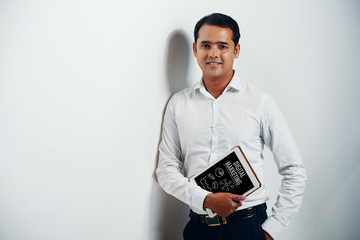 Elegant Indian digital marketing professional leaning on white wall with tablet computer in his hand