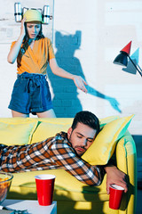 Wall Mural - shocked girl in beer hat looking at drunk man sleeping on couch after home party