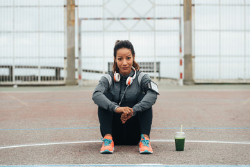 Wall Mural - Relaxed fitness woman drinking detox green smoothie during outdoor city workout rest. Sport nutrition and healthy lifestyle concept. Female athlete taking a break sitting on the ground.