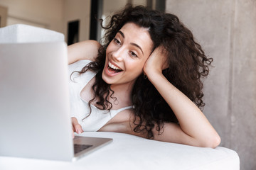 Wall Mural - Photo closeup of young adorable woman with long curly hair wearing silk leisure clothing lying on couch in apartment, and laughing while looking at screen of laptop