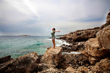 Fototapeta Natura - Woman with outstretched arms enjoying the wind and breathing fresh air on the rocky beach 