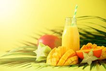 Delicious Juicy Smoothie With Orange Fruit And Mango On Yellow Background. Copy Space. Pop Art Design, Creative Summer Concept. Fresh Juice In Glass Bottles Over Green Palm Leaves. Banner