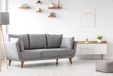 Fototapeta  - Simple, gray sofa standing next to a white cupboard in living room interior with decorations on wooden shelves. Real photo
