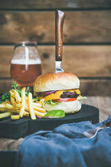 Wall Mural - Classic burger dinner. Beef meat homemade burger with glass of beer, French fries and salad on dark wooden serving board, rustic wooden wall at background, copy space, vertical composition