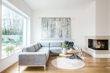 Two hairpin tables with books, tulips and coffee cup placed on white rug in bright living room interior with grey sofa, fireplace and abstract painting on the wall