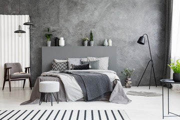 grey armchair and stool near bed with headboard in bedroom interior with black lamp. real photo