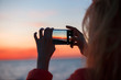 © Maksim Kostenko - Woman traveler using smartphone and taking photo of colorful sea sunset