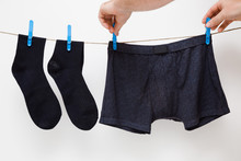 Man's Hands Hanging Clean Black Socks And Boxer Shorts. Underwear Drying On Laundry Rope At White Wall. Dry Cleaning. Regular Washing. Daily Routine. Clothes Care.