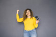 Cute brunette plus size woman with curly hair in yellow sweater and jeans standing on a neutral grey background. She holds a joystick, playing on console games