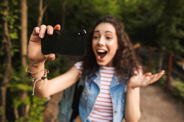 Wall Mural - Blurry photo of excited joyful woman 18-20 with backpack, shouting and taking selfie photo on focused black cell phone while walking in green park