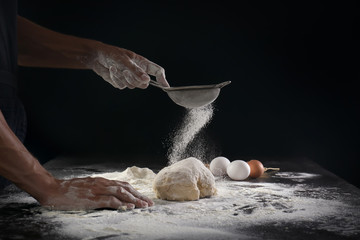 Man sifting flour over dough on black background