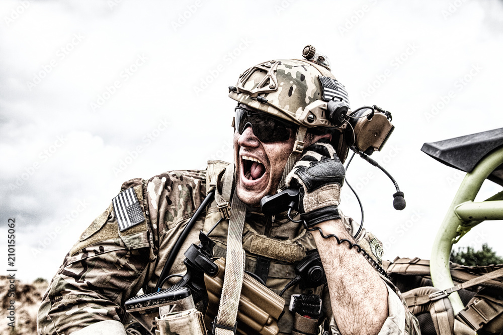 Special forces soldier, military communications operator or maintainer in helmet and glasses, screaming in radio during battle in desert. Calling up reinforcements, reporting situation on battlefield - obrazy, fototapety, plakaty 