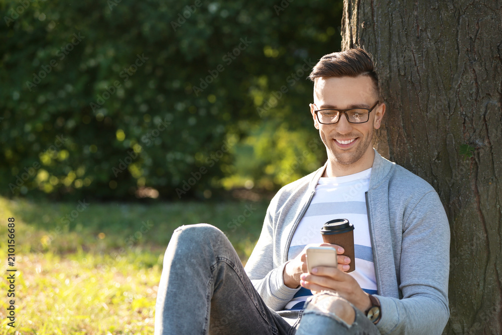 Handsome man with mobile phone and cup of coffee resting in park