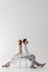 Wall Mural - side view of young couple of models in linen clothes sitting back to back on chair isolated on grey background