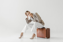 Young Stylish Female Model In Linen Jacket Sitting On Vintage Suitcase And Looking At Camera Isolated On Grey Background