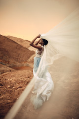 Wall Mural - Black bride stands and holds waving bridal veil in her hands on background of beautiful landscape.