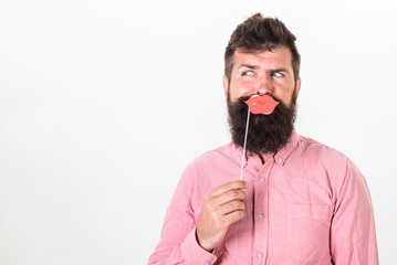 Wall Mural - Man with trendy beard holding paper lips. Bearded man making funny faces isolated on white background. Hipster with long beard in pink shirt posing with paper accessories, party memories concept